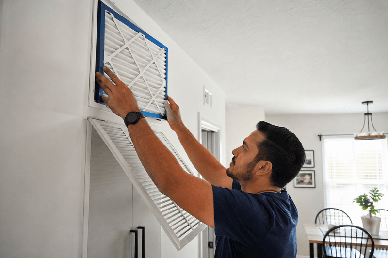 Homeowner sliding a new MERV 13 pleated air filter into a hallway return vent. 