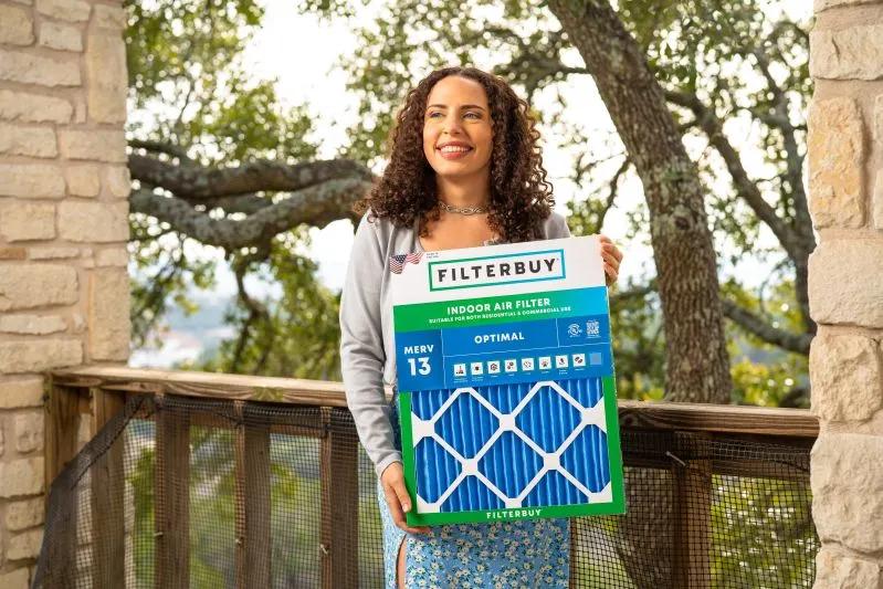 Smiling woman holding a large Filterbuy 9x30x1 MERV 13 indoor air filter box on a balcony surrounded by trees