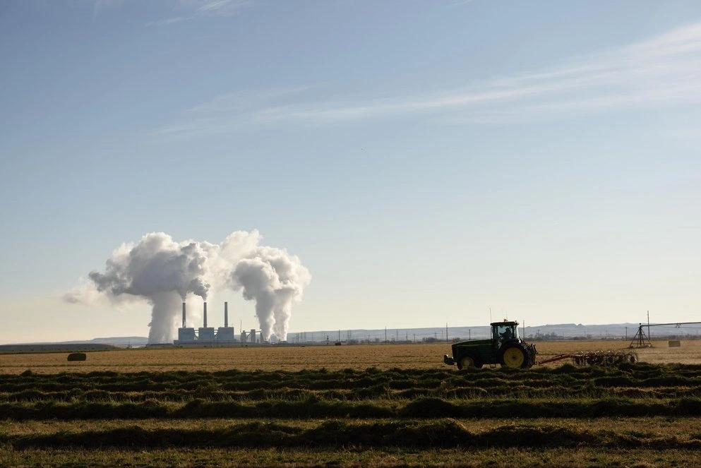States and Counties With the Highest Rates of Lung Cancer - Infographics of a tractor working near a coal power plant in an open field, reflecting the impact of rural industrial emissions in states like Wyoming.