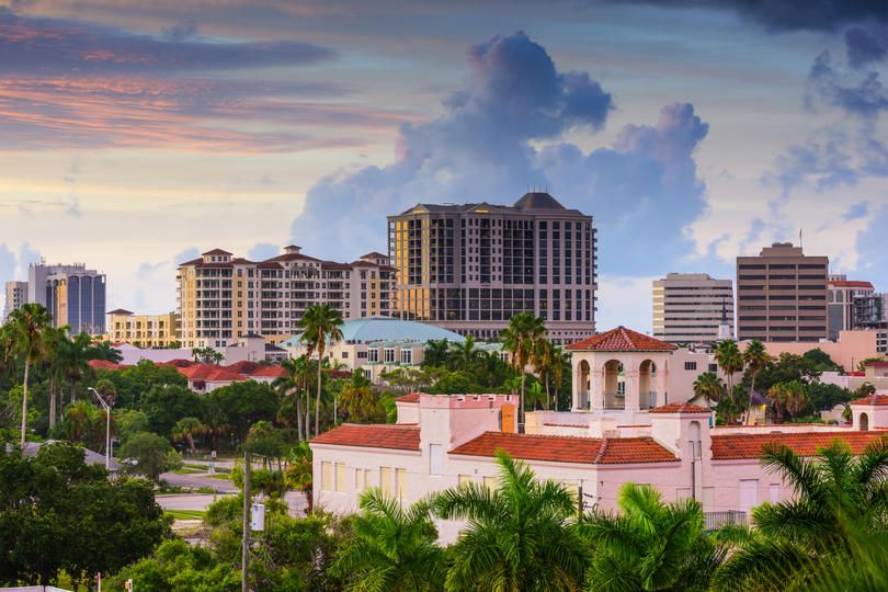 A view of a city skyline with modern buildings and palm trees, set against a sky with soft clouds during sunset or twilight. The scene reflects a blend of urban development and tropical surroundings.