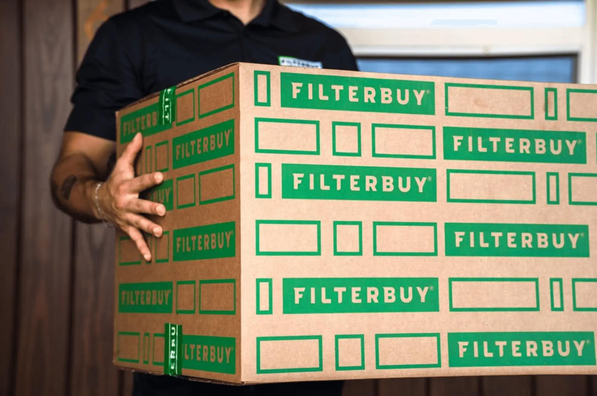 An image of a delivery worker carrying a large stacked Filterbuy shipping box labeled with green branding.