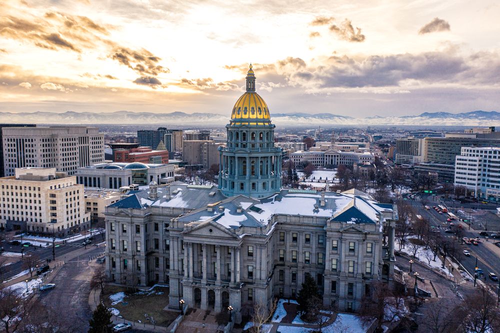 Historic government building with a gold dome surrounded by snow-covered rooftops and distant mountains at sunset.