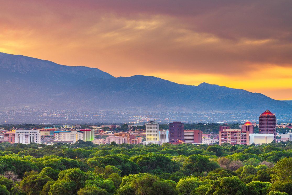 A view of a city at sunset, with the skyline and buildings in the foreground, framed by lush greenery and mountains in the background, under a colorful sky.