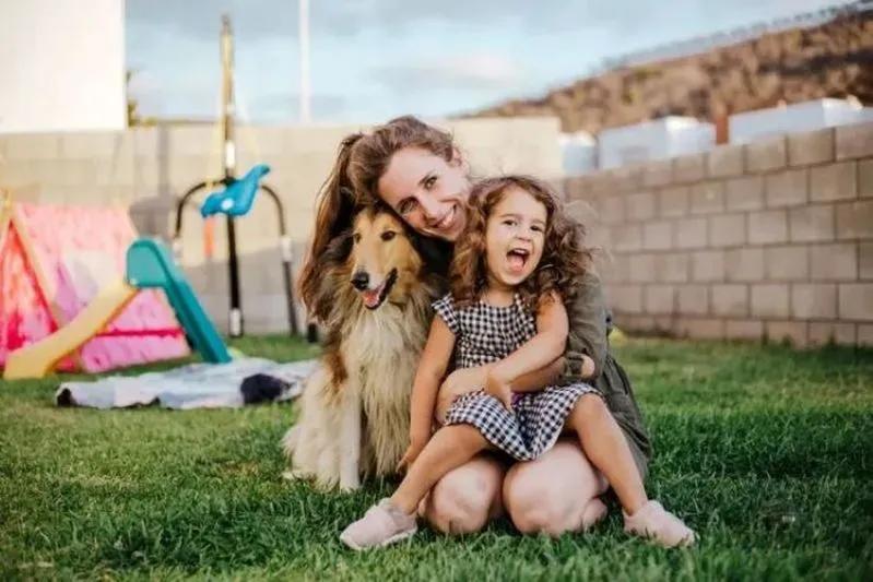 View of a happy mother and daughter playing with Collie dog in backyard with playground in the background