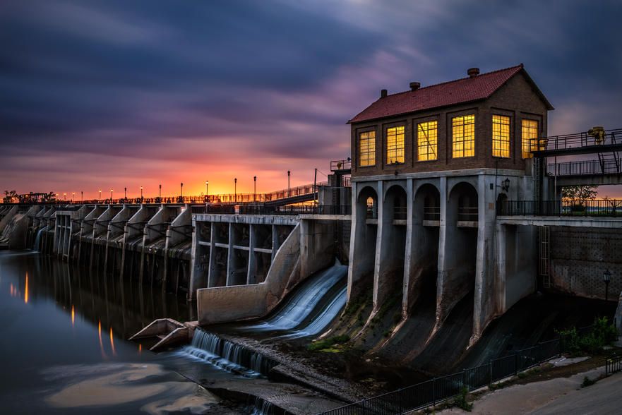 A view of a dam at sunset, with water flowing over the spillways and lights glowing from a building at the structure, set against a dramatic, cloudy sky.