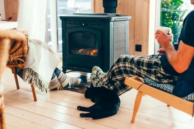 A view of a cozy indoor scene with people relaxing by a wood-burning stove, wrapped in plaid blankets, with a black cat lying on the floor.