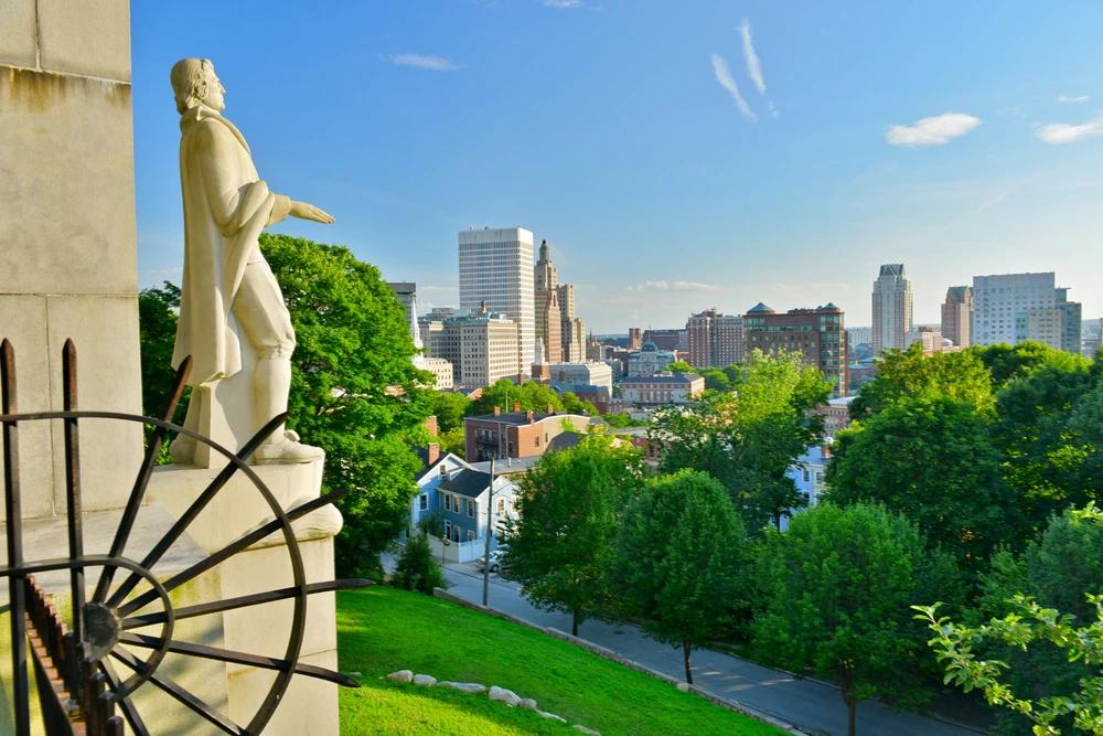 Statue overlooking downtown Providence, Rhode Island with historic architecture, city skyline, and lush summer greenery.