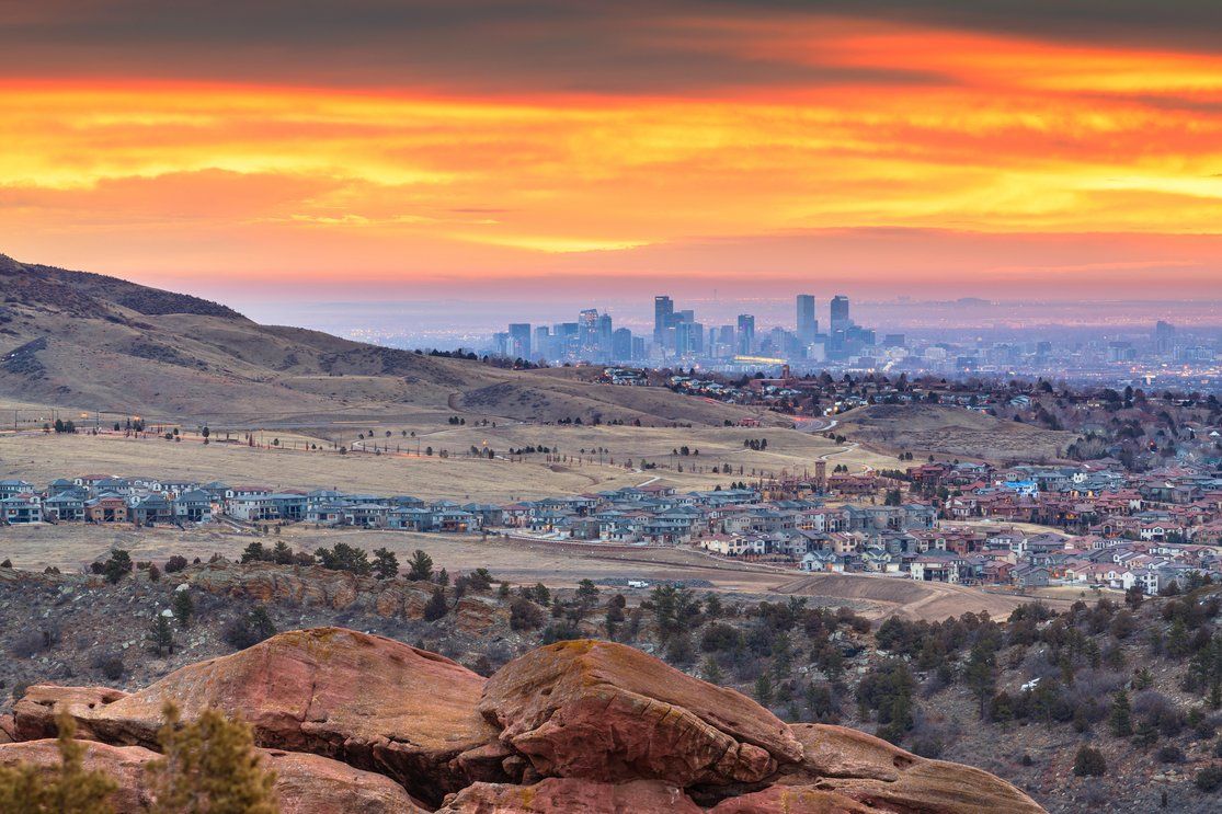 A view of a city skyline at sunset, with rolling hills and homes in the foreground, and the vibrant orange and purple sky setting the scene behind the urban landscape.