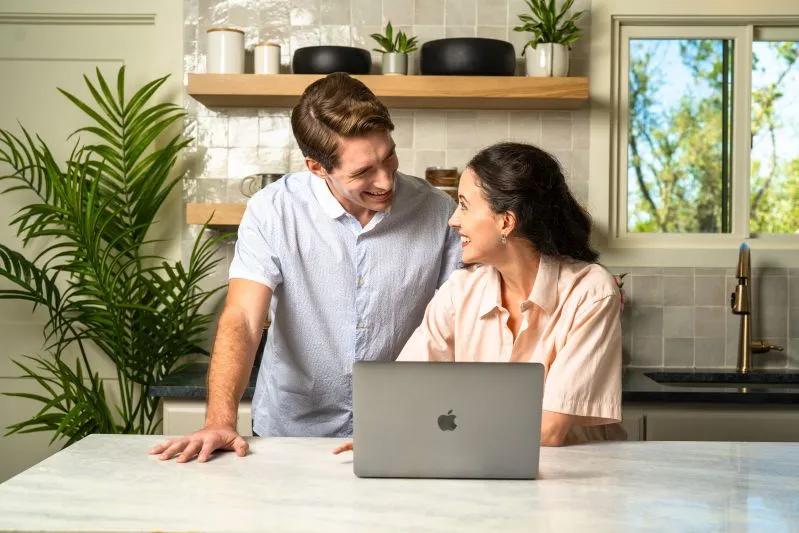 View of smiling couple using laptop together in modern kitchen with natural light and indoor plants