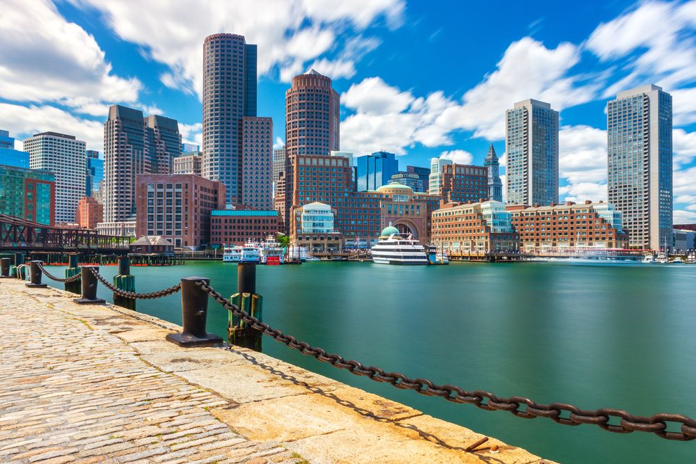 City skyline seen from a cobblestone pier, with tall buildings, boats, and calm waterfront on a partly cloudy day.