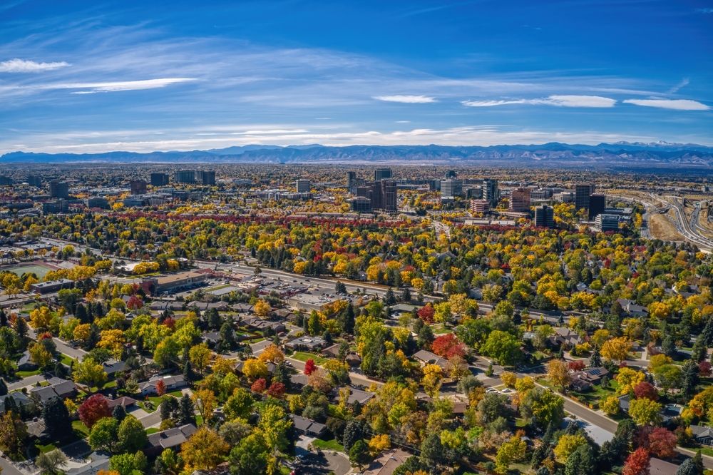 An aerial view of a city surrounded by colorful autumn trees, with vibrant shades of red, orange, and yellow blending with the urban landscape and distant mountains under a clear blue sky.