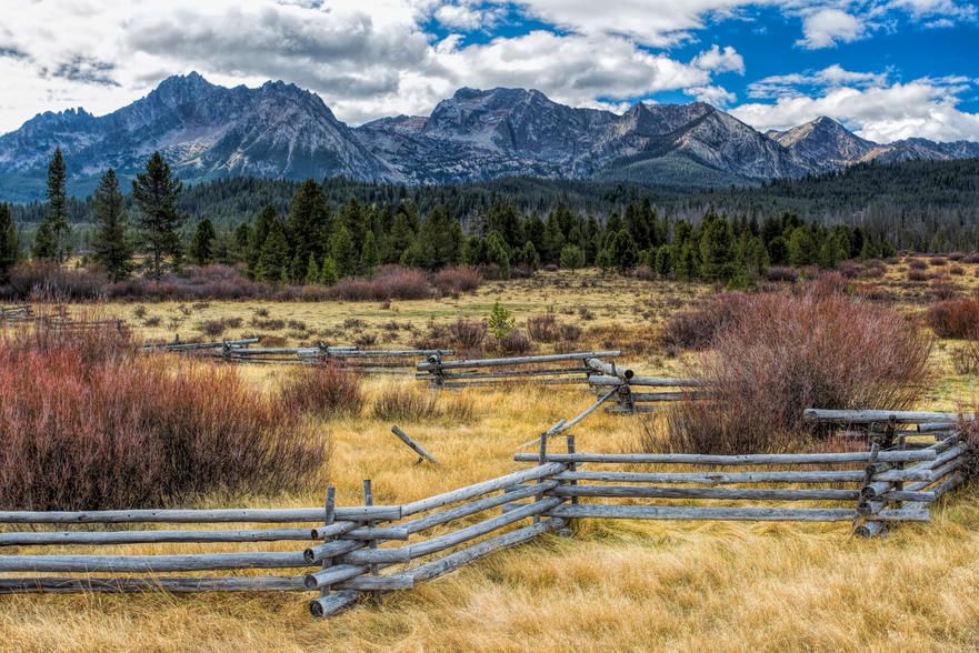A view of a vast open field with rustic wooden fences, set against a backdrop of snow-capped mountains, lush forest, and partly cloudy skies. The scene conveys a serene and expansive natural landscape.
