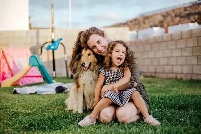 A view of a joyful mother, daughter, and dog sitting on the grass in a backyard, enjoying clean outdoor air and family time.