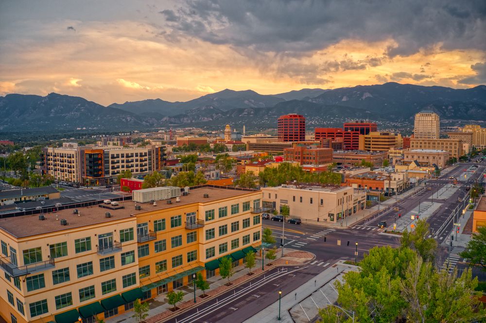 A panoramic view of a city with modern buildings and wide streets, set against a backdrop of mountains. The scene is bathed in warm light during sunset, highlighting the urban landscape.