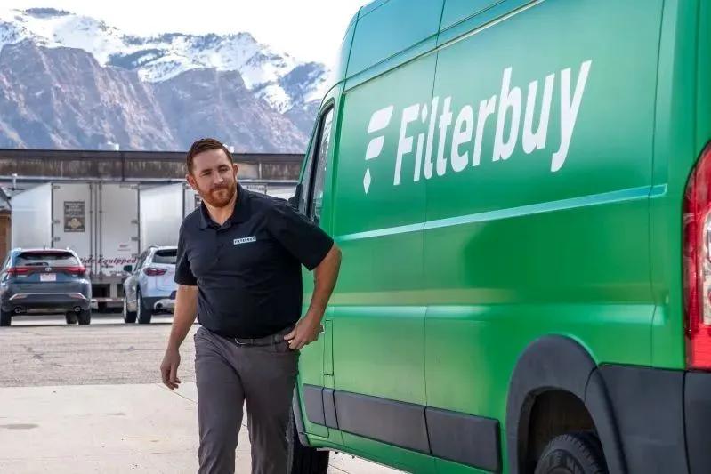 A view of a Filterbuy delivery van with a uniformed employee standing beside it, set against a backdrop of snow-capped mountains.