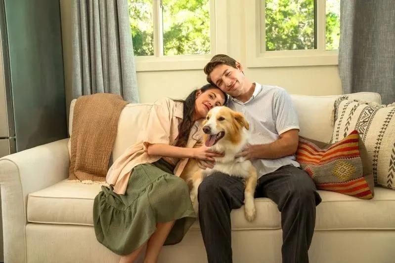A view of a happy couple sitting on a couch with their dog in a bright, cozy living room.