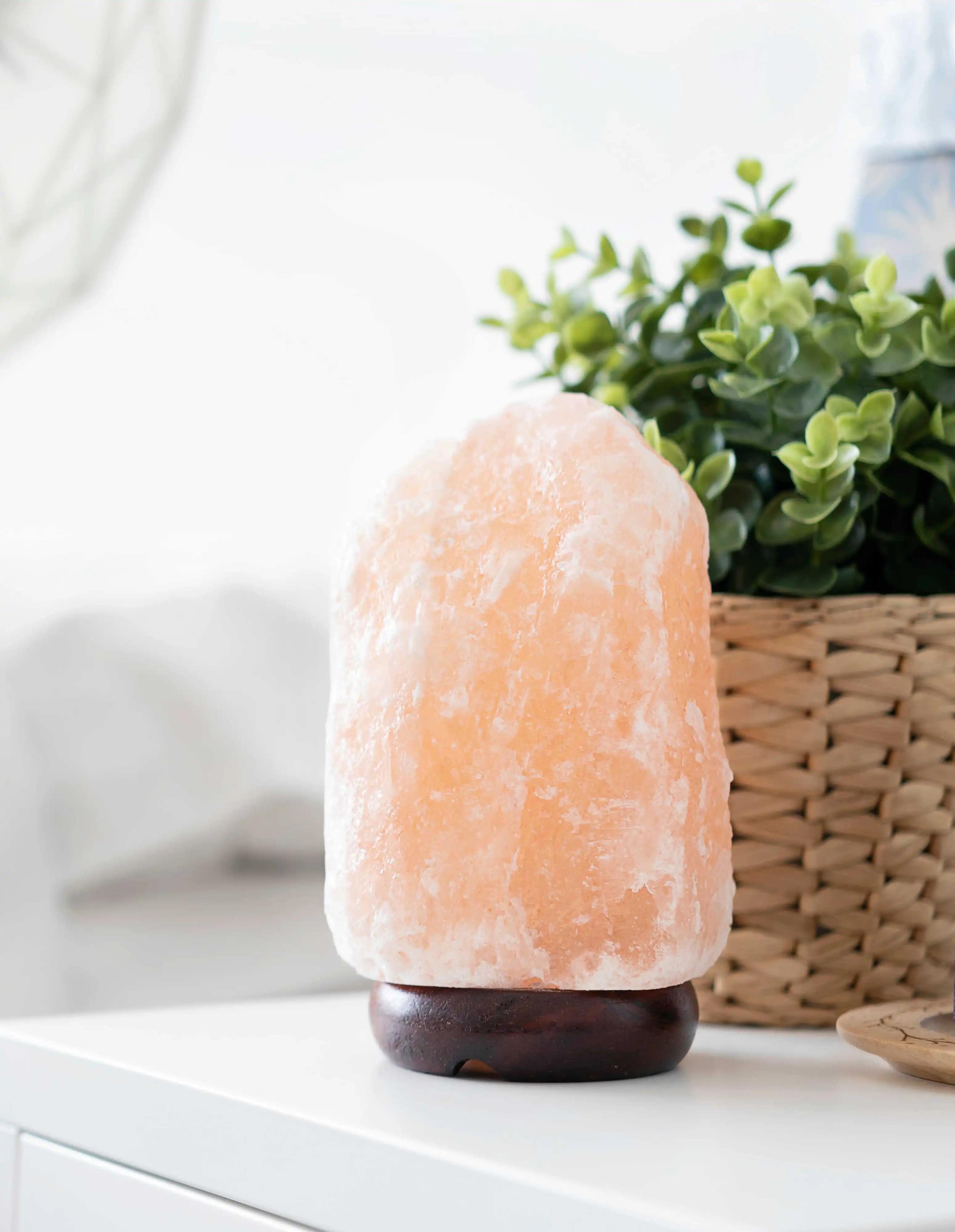 View of a pink Himalayan salt lamp on a white table next to a green potted plant in a woven basket. 