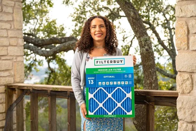 View of a smiling woman holding a Filterbuy indoor air filter on an outdoor balcony, with trees in the background.