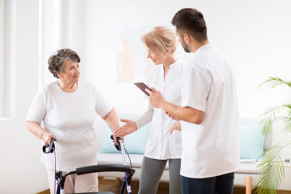 Elderly woman with a walker receiving assistance from two healthcare professionals in a clinical setting.