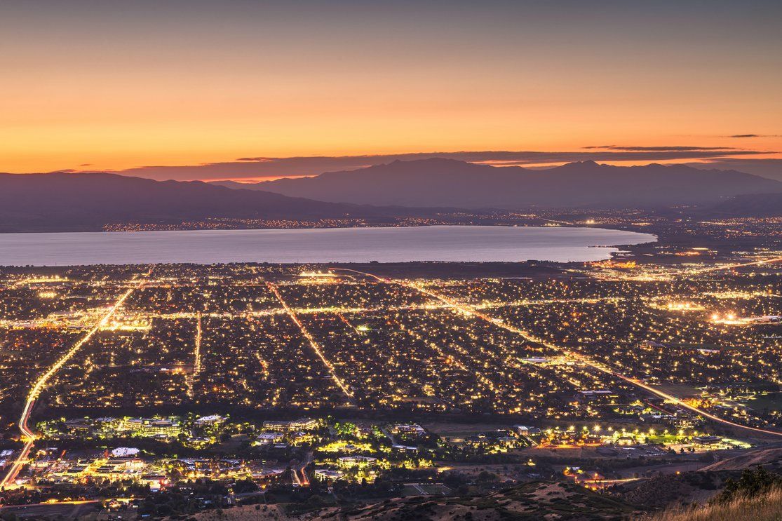 A view of a city illuminated at night with streetlights and buildings glowing, set against the backdrop of a calm body of water and distant mountains under a colorful sunset sky.