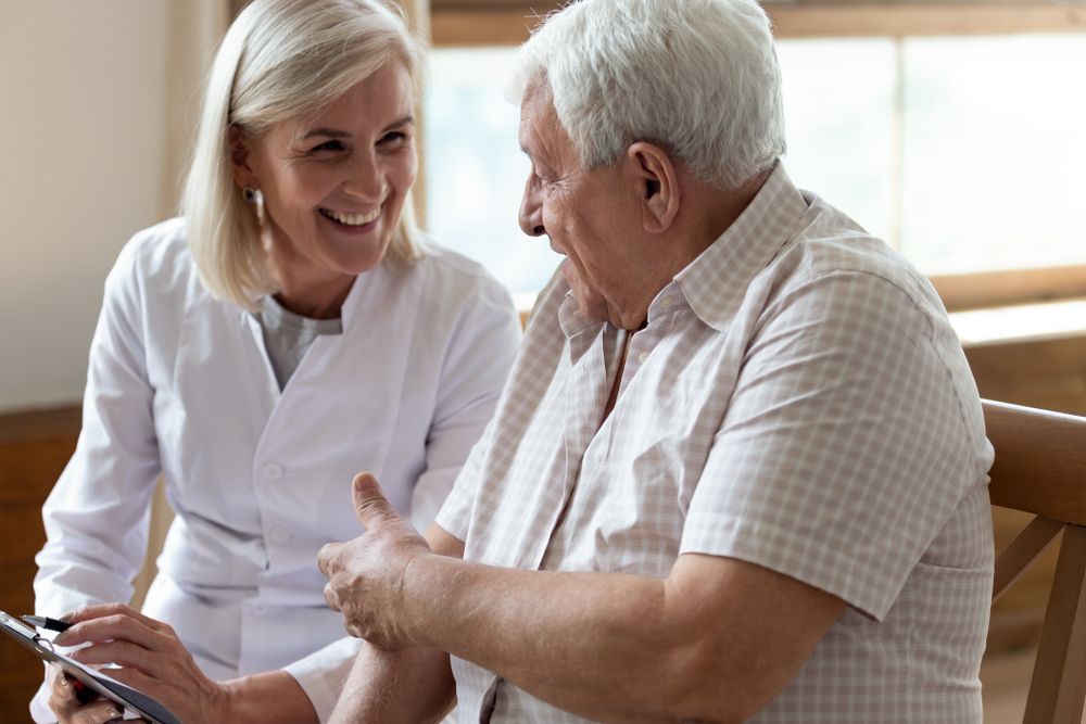 Smiling female healthcare worker talking to an older male patient while holding a clipboard.