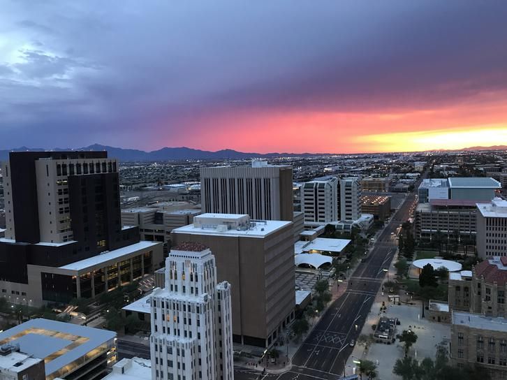 A view of a city at sunset, with tall buildings and streets visible, and a colorful sky with vibrant hues of orange, purple, and pink in the distance behind the mountains.