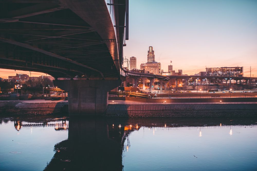 Omaha, Nebraska skyline at sunset viewed from beneath a bridge, with reflections in the river.