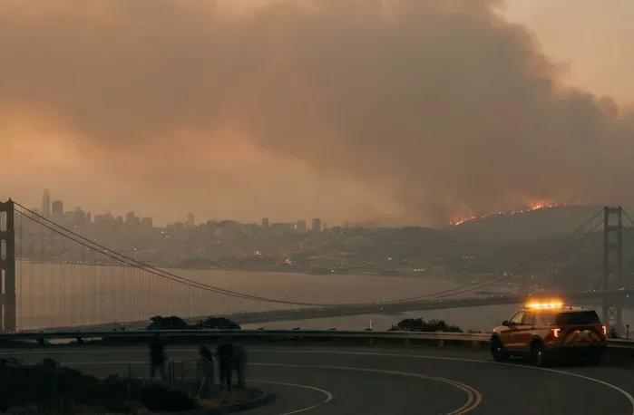 Wildfire smoke and flames fill the sky over San Francisco as seen from the Marin Headlands with the Golden Gate Bridge in the foreground and an emergency response vehicle with flashing lights parked on the roadside while onlookers watch the fire burn across the hillside during a Northern California wildfire event