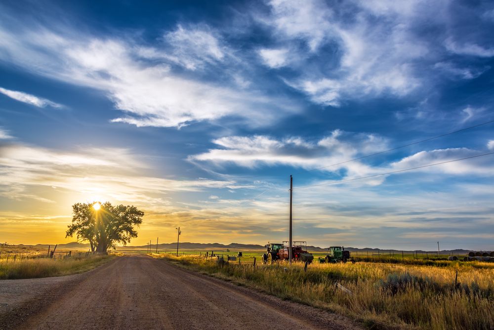 Country road leading toward the sunset, flanked by grassy fields and farming equipment beneath a vivid, expansive sky.