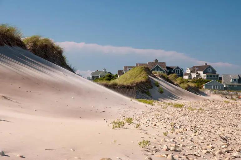 View of coastal sand dunes with scattered vegetation and beachfront houses under a clear blue sky, capturing a serene shoreline landscape.