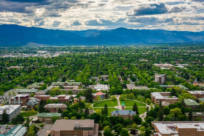A view of a green, tree-filled city with buildings and open spaces, set against a backdrop of mountains under a partly cloudy sky. The area appears to be well-developed with a focus on greenery and urban planning.