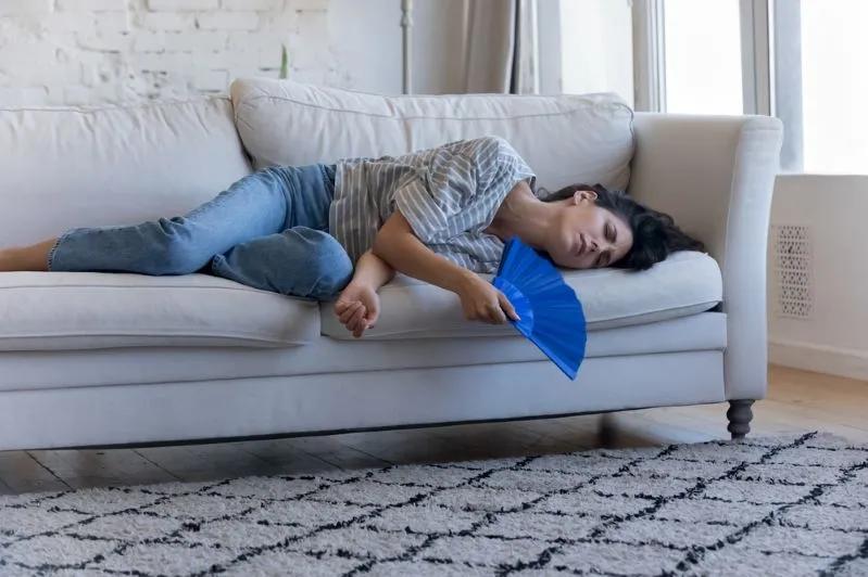 Image of exhausted woman lying on couch fanning herself due to heat inside home without air conditioning.