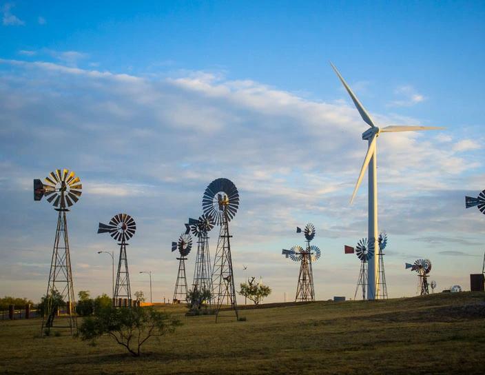 View of various windmills and a modern wind turbine on an open field under a blue sky, showcasing traditional and renewable energy side by side.