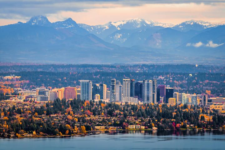 A view of a city skyline with tall buildings, surrounded by colorful autumn trees, set against a backdrop of snow-capped mountains and a calm lake.