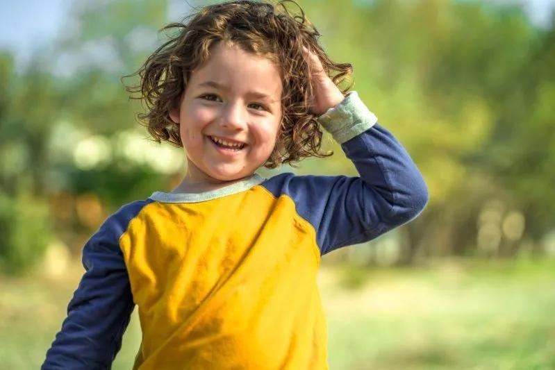 A view of a cheerful young child with curly hair smiling outdoors, enjoying a sunny day in nature.