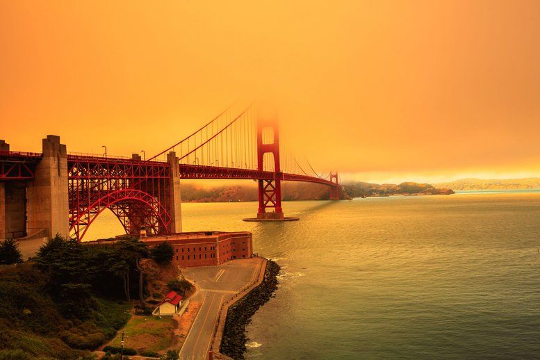 A view of the Golden Gate Bridge partially shrouded in smoke, with an orange-hued sky and calm waters below, possibly due to nearby wildfires affecting the atmosphere.