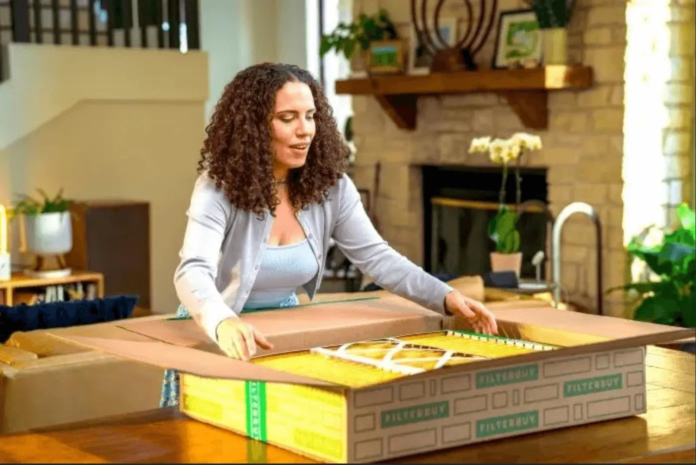 An image of a woman opening a large Filterbuy box filled with air filters inside a cozy living room.