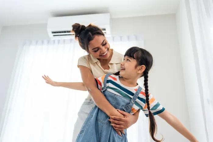 A view of a young girl being playfully lifted by her mother in a bright, airy room with an air conditioning unit overhead, illustrating the benefits of using effective air filters.