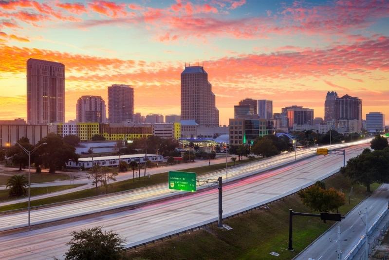Cities With the Most Multigenerational Households - Image of Orlando city skyline at sunrise, showcasing tall buildings with traffic moving on highways, and a colorful sky in the background.