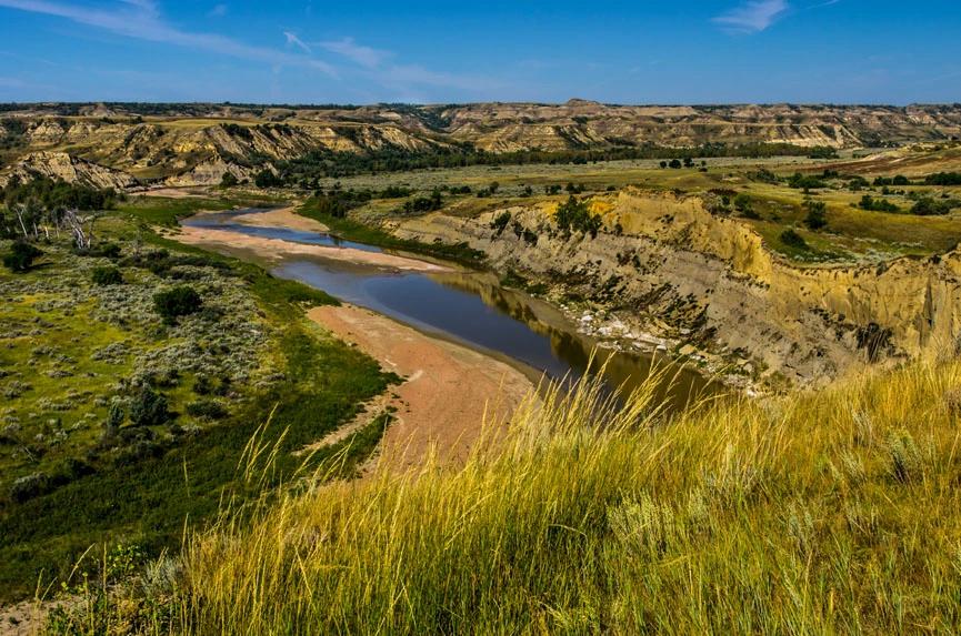 View of a winding river cutting through a rugged valley with grassy hills and eroded cliffs under a clear blue sky.