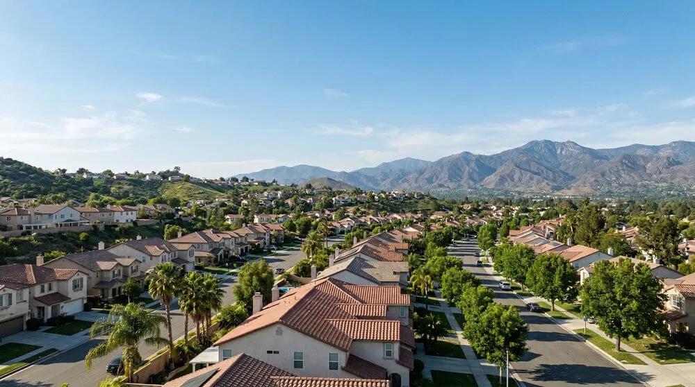 An image of a sunny suburban neighborhood in Riverside, California, featuring neatly organized houses with terracotta roofs, palm trees, and a distant mountain range.