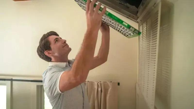 A view of a man installing a new air filter into a ceiling vent, demonstrating routine HVAC maintenance in a home setting.