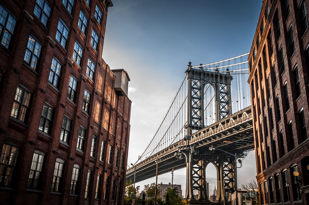 Steel suspension bridge framed between two old brick buildings in a cityscape.