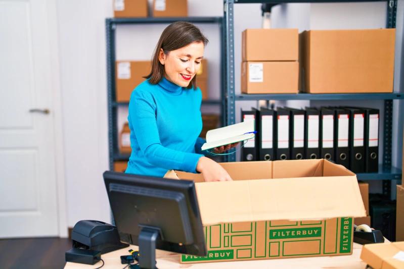 View of - A woman in a blue sweater packs items into a Filterbuy box in a shipping office. 