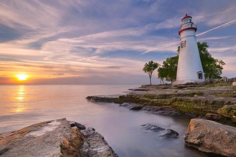 View of a lighthouse on a rocky shoreline at sunset, with calm waters and pastel skies reflecting a tranquil scene.