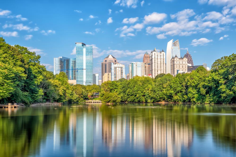 Atlanta skyline reflected in a serene lake surrounded by lush green trees under a partly cloudy blue sky.