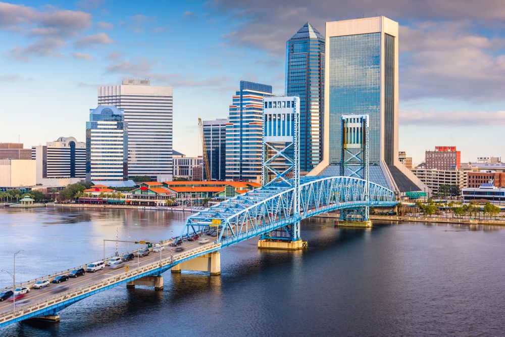 A view of Jacksonville's skyline with the blue Green Monster Bridge crossing the St. Johns River, surrounded by modern buildings and cityscapes under a bright sky.