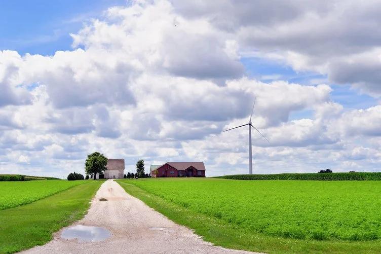 View of a rural farmstead with a gravel road, green fields, and a wind turbine under a bright, cloudy sky.